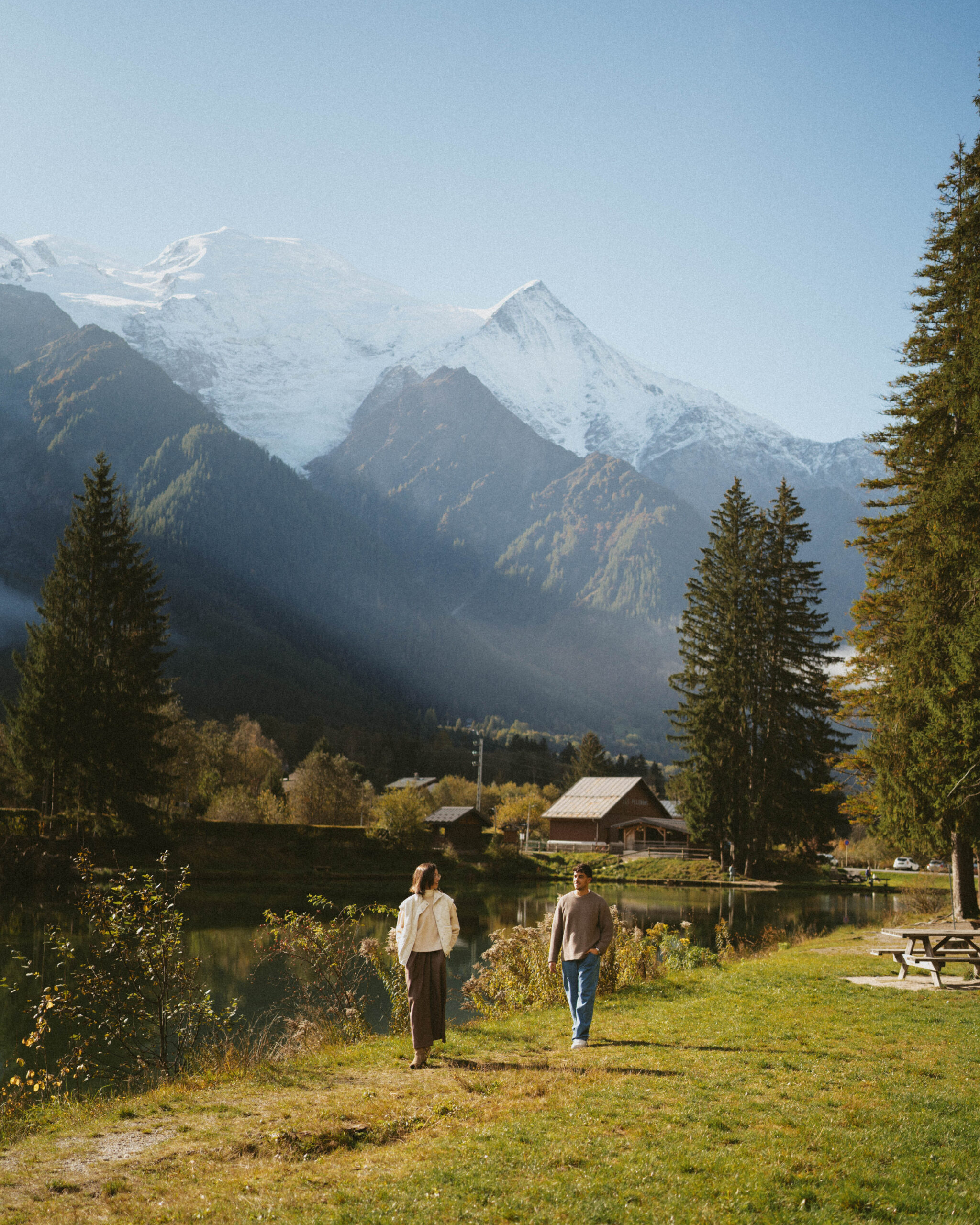 Katya and Michael walking along a lakeside path surrounded by lush greenery, with the majestic French Alps and snow-capped peaks in the background during their elopement.